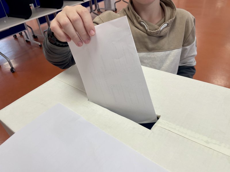 Ein Kind steckt einen weißen Umschlag in eine Wahlurne aus Pappe. Die Umgebung zeigt einen Raum mit Stühlen.