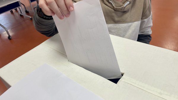 Ein Kind steckt einen weißen Umschlag in eine Wahlurne aus Pappe. Die Umgebung zeigt einen Raum mit Stühlen.