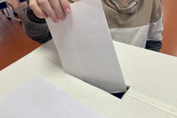 Ein Kind steckt einen weißen Umschlag in eine Wahlurne aus Pappe. Die Umgebung zeigt einen Raum mit Stühlen.