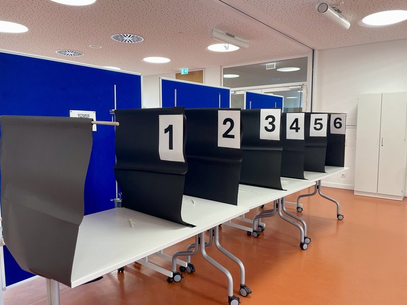Sechs Wahlurnen mit schwarzen Sichtschutzwänden stehen auf einem Tisch in einem hellen Raum.